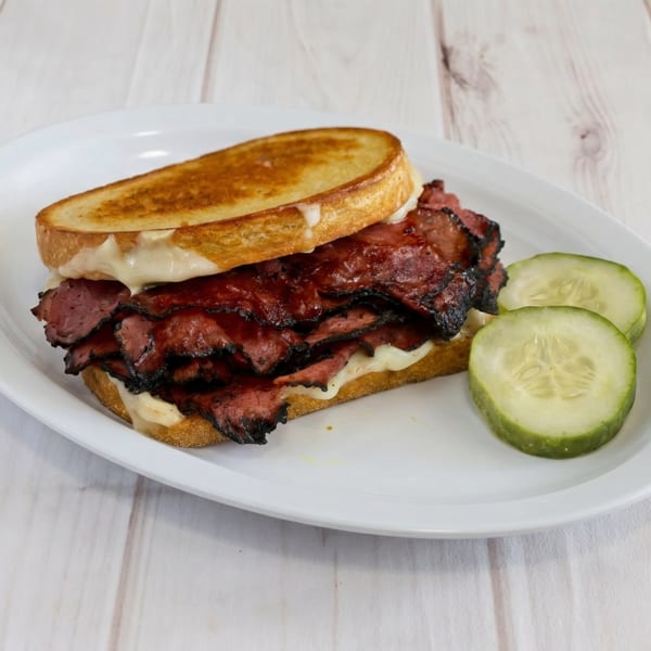 A grilled pastrami sandwich with melted cheese and two cucumber slices on a white plate.