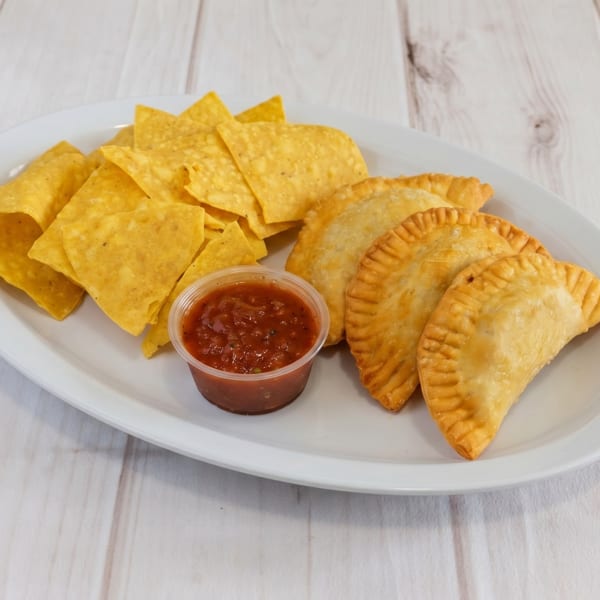 A white platter with golden empanadas, tortilla chips, and a cup of salsa on a wooden table.
