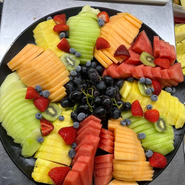 A large black platter filled with an assortment of sliced fresh fruits including melon, pineapple, and berries.