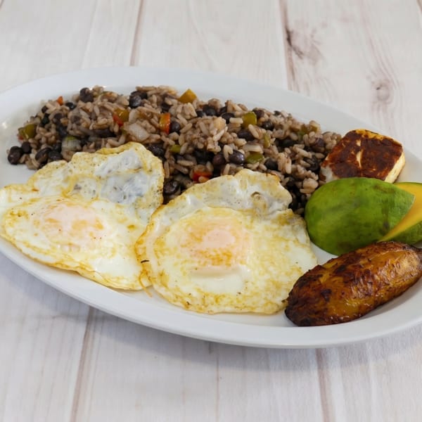Gallo pinto with two fried eggs, fried plantains, avocado slices, and fried cheese on a white plate.
