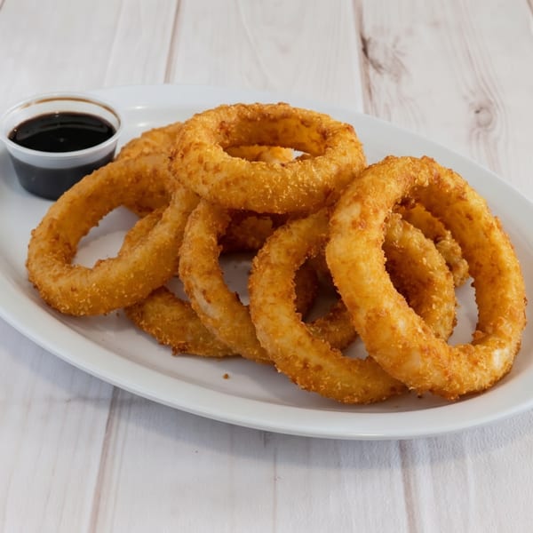 Golden-brown onion rings piled on a white plate with a side of dark dipping sauce.